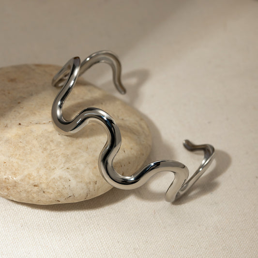High-polish stainless steel sculpted wave cuff bracelet with minimalist open design displayed on a beige stone, showcasing its sleek and modern design.