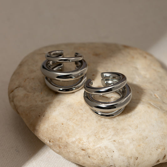 Sculpted layered stainless steel cuff earrings with polished silver-tone finish displayed on a beige stone.