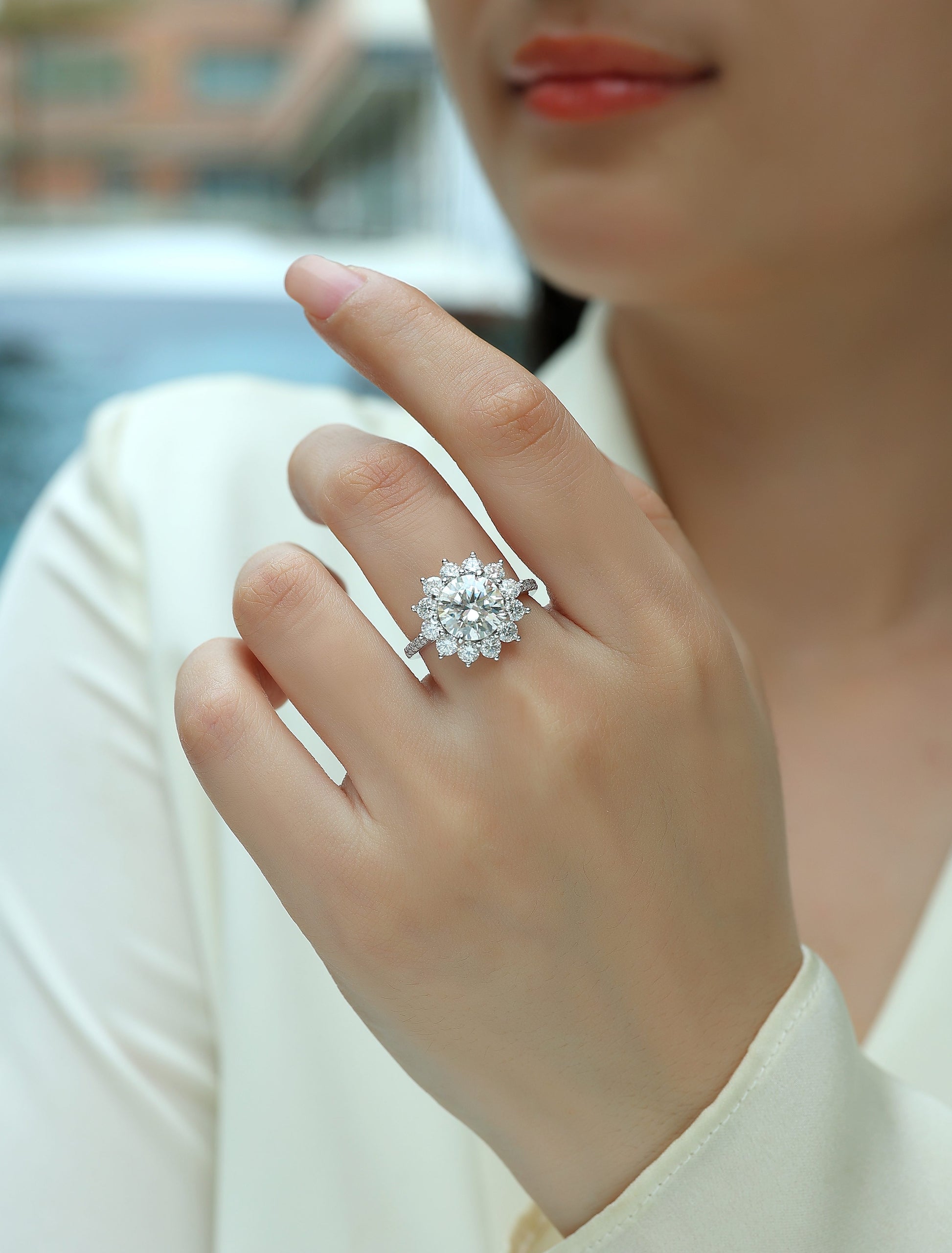 Close-up of a hand wearing a Soléa 3 carat moissanite floral halo ring in platinum-plated sterling silver statement engagement ring by MerchLuxx with a blurred background