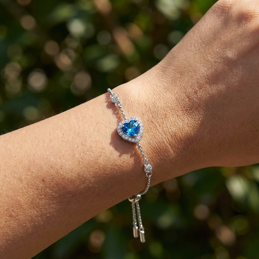 A Celestara blue moissanite heart bracelet in platinum-plated sterling silver with heart accents by MerchLuxxon against a blurred green background