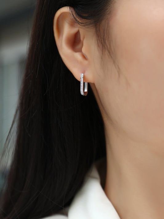 Close-up of an ear wearing the Avelisse pavé moissanite elongated huggie hoop earrings in platinum-plated sterling silver by MerchLuxx  with a blurred background