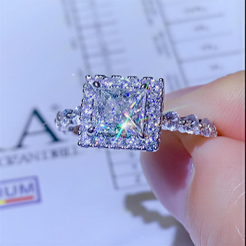 1 carat square-cut moissanite halo ring in platinum-plated sterling silver with pavé side stones held by a hand against a blurred background